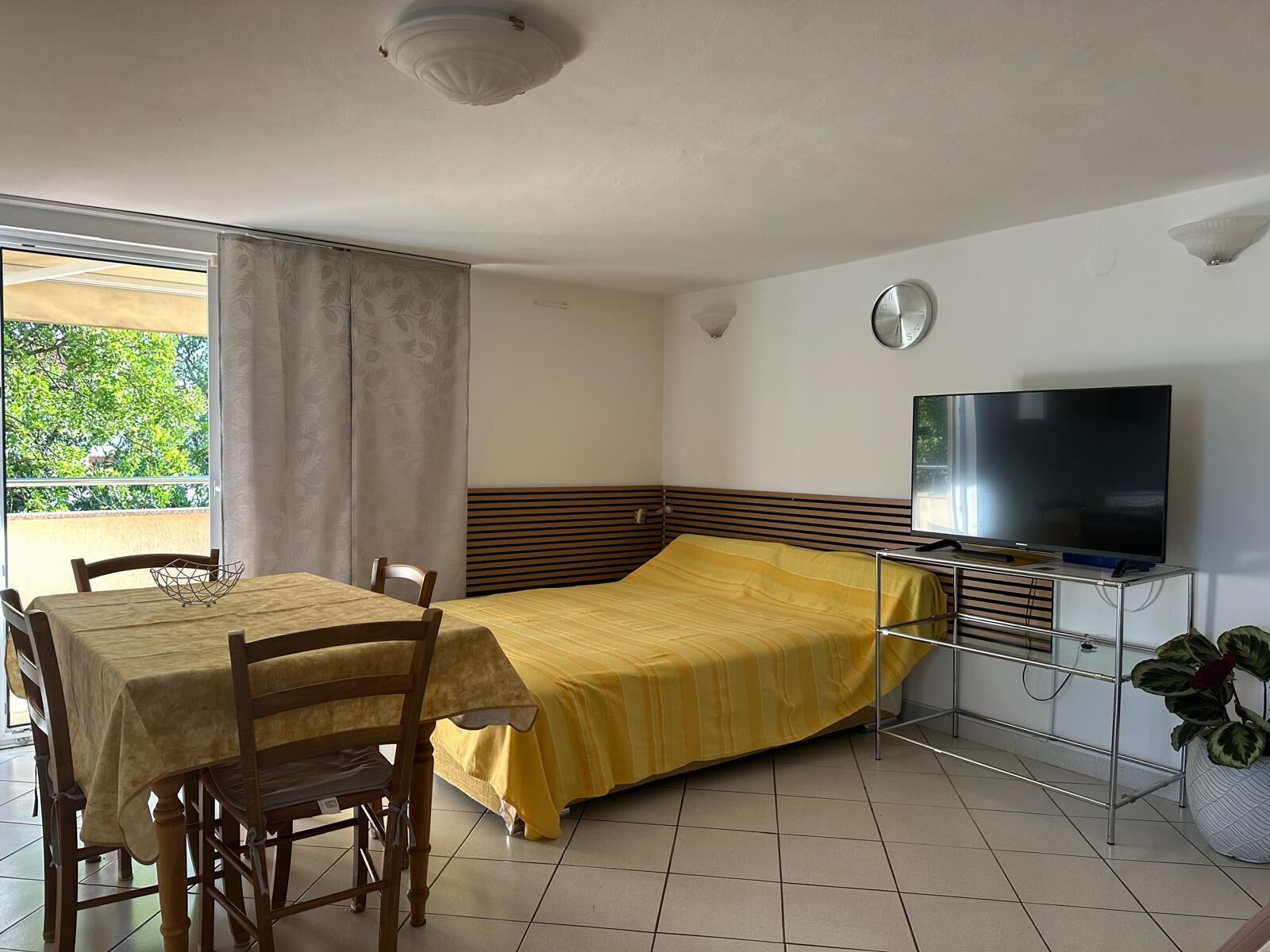 A room with a bed, a TV, a dining table with chairs, and a window with curtains.