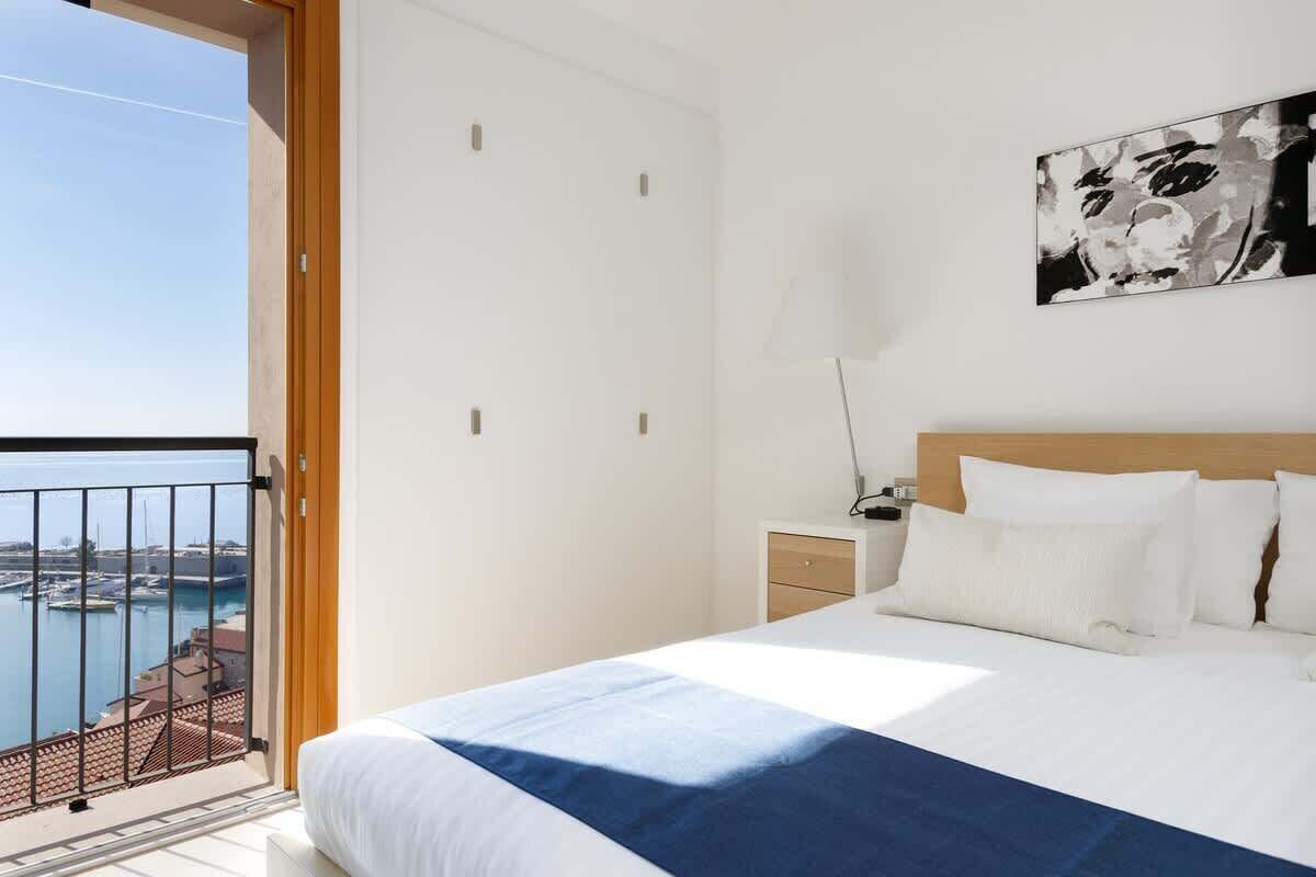 A bedroom with a bed, a nightstand, a lamp, a painting, and a balcony with a view of the water.
