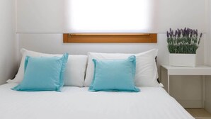 Un lit avec une literie blanche et deux oreillers bleu clair, une tête de lit en bois, une table de chevet blanche ornée d’un pot de lavande et une fenêtre avec des stores.