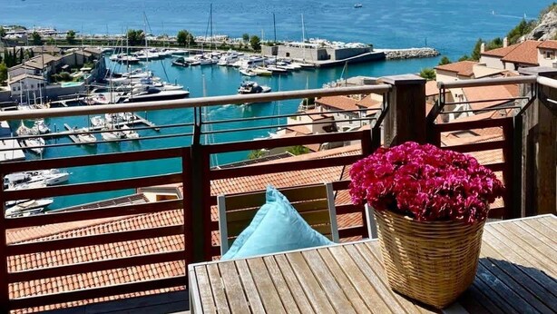 Un balcon donnant sur un port de plaisance, une table en bois et un panier en osier rempli de fleurs.