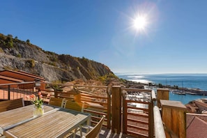 A wooden deck with tables and chairs overlooking a coastal area with a clear view of the sea and a rocky hillside.