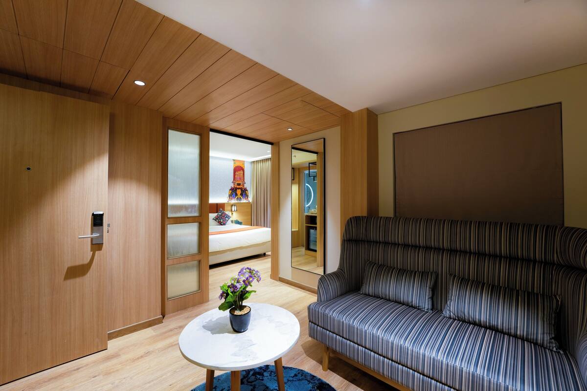 A modern hotel room with a wooden ceiling, a striped sofa, a round table with a plant, and a bed with a curtain.