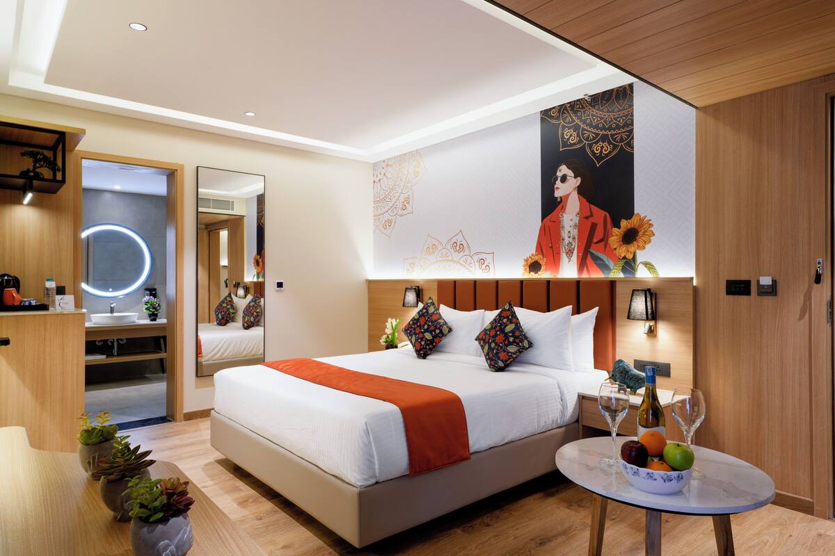 A modern hotel room with a large bed, bedside table with wine glasses, a mirror, and a wall with a poster.