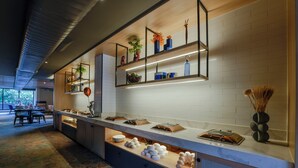 A modern hotel kitchen with a long counter, shelves with plants, and a dining area in the background. - TGI Oberon  (Bengaluru)