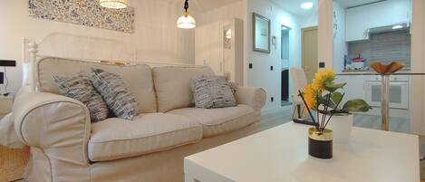 A living room with a beige sofa, a white coffee table, and a vase with flowers. There is a kitchen visible in the background.