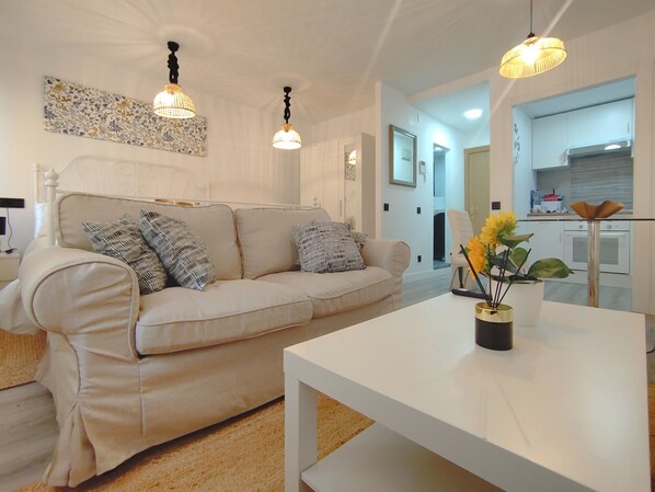 A living room with a beige sofa, a white coffee table, and a vase with flowers. There is a kitchen visible in the background.