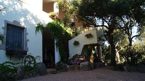 A building with a stone facade, a window with a metal grill, and a door. There are plants and flowers around the entrance. - Agriturismo Le Sughere (Padru)