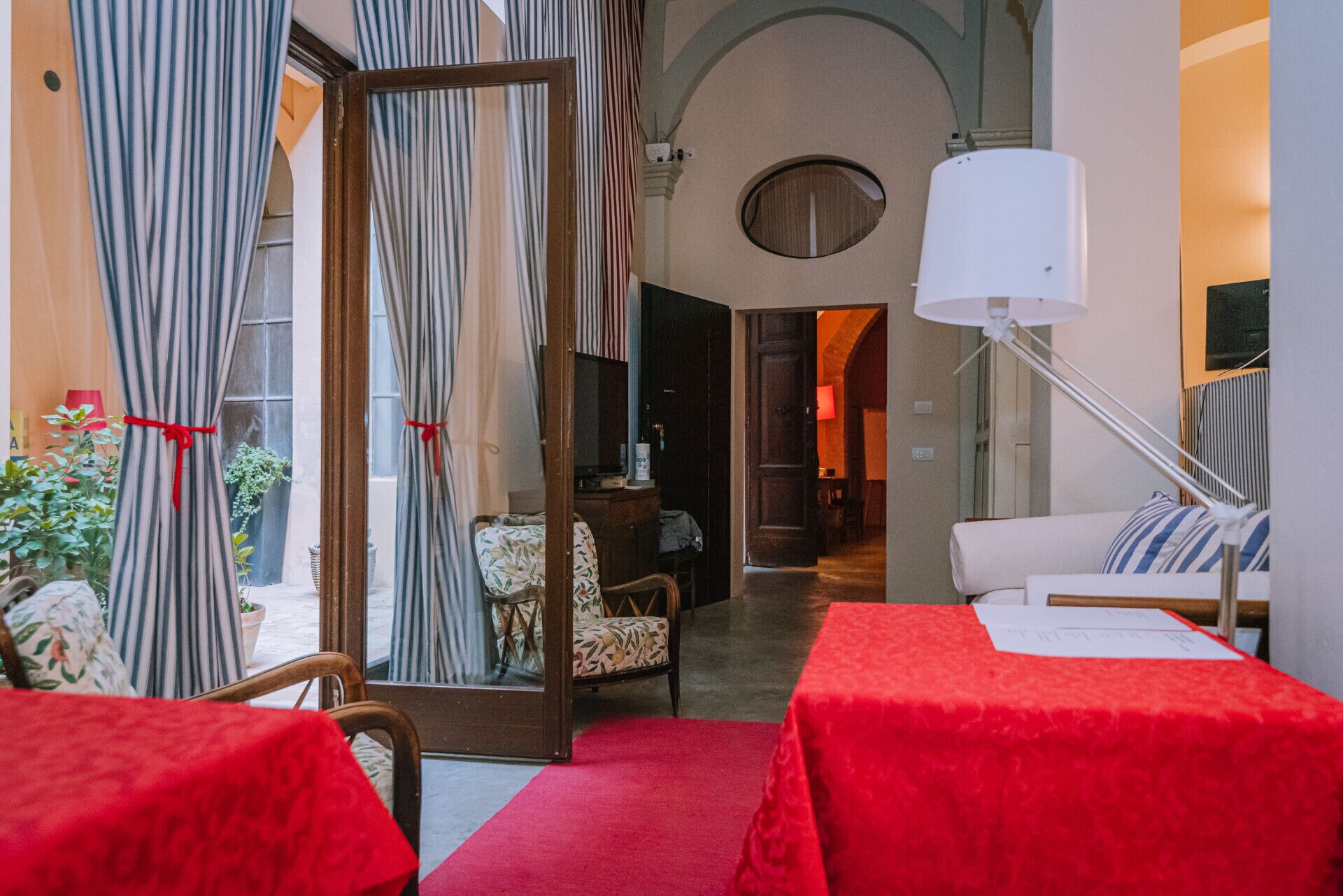 Une chambre d’hôtel avec un tapis rouge, un canapé blanc, une table recouverte d’une nappe rouge, des rideaux à rayures et une lampe.
