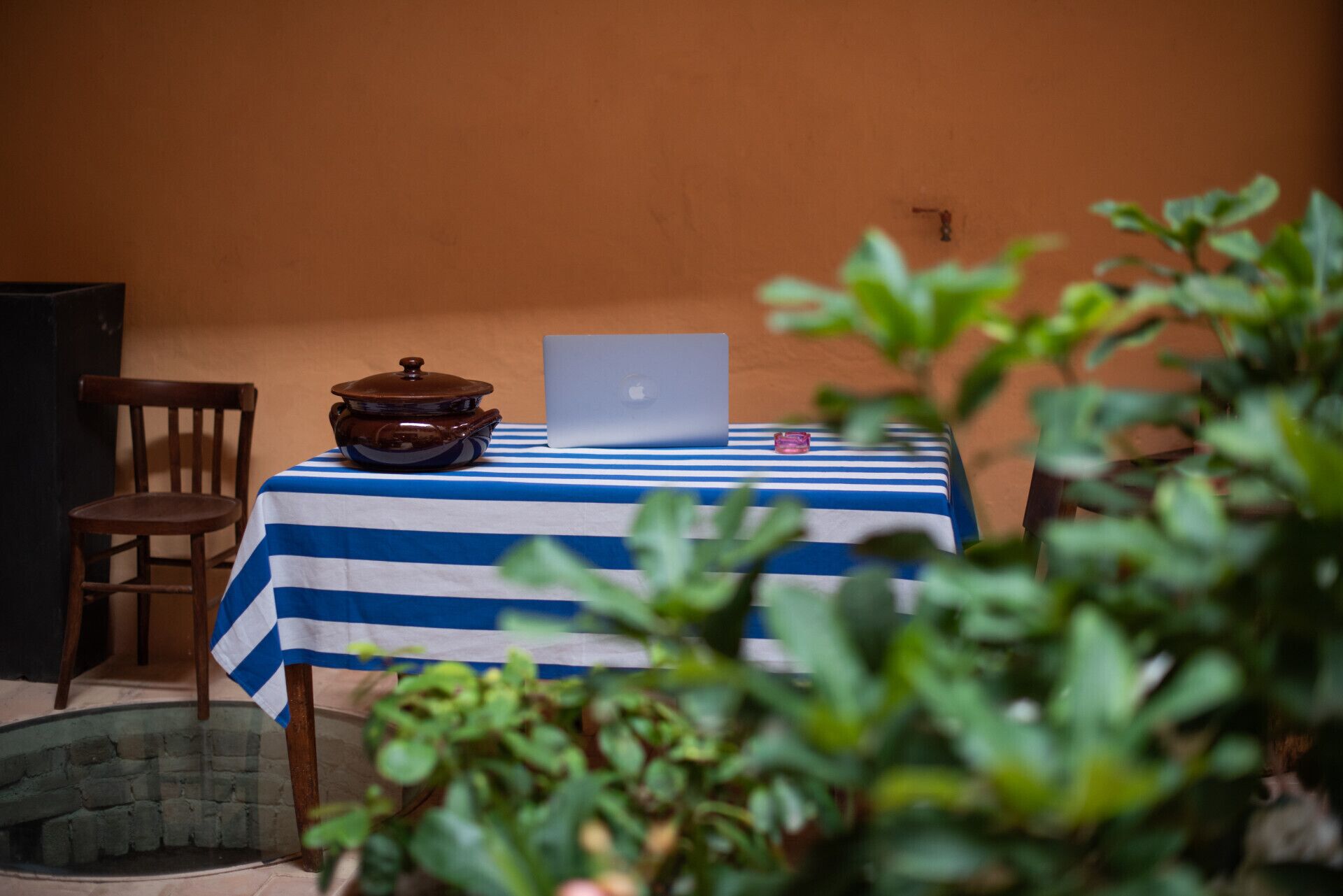 A table with a laptop and a pot on a striped tablecloth, a chair, and a wall in the background.