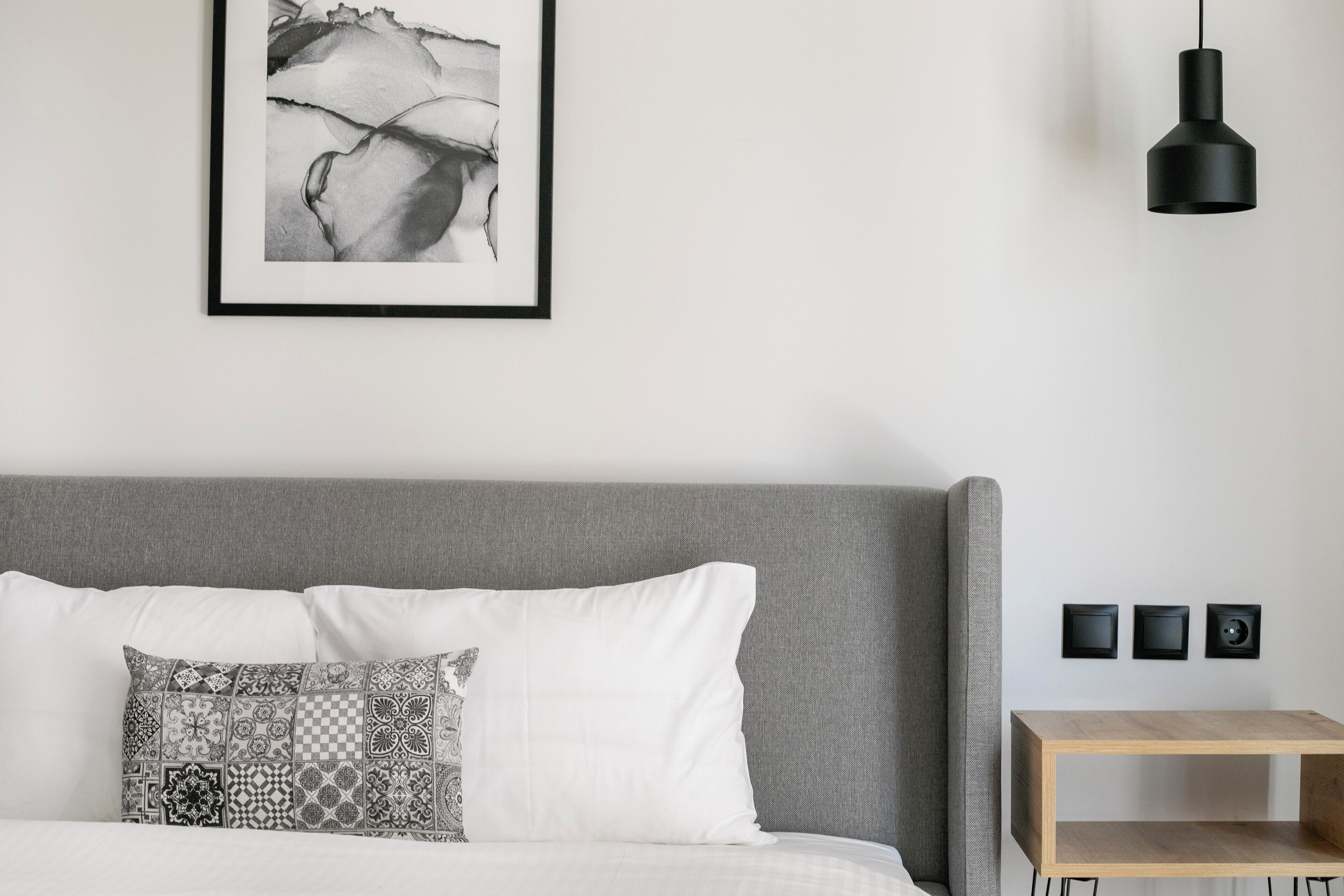 A modern bedroom with a grey headboard, a white bed with patterned pillows, a black pendant light, and a small wooden nightstand with three black speakers.