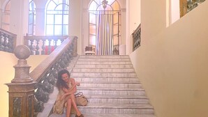 Eine imposante Treppe mit Marmorstufen und aufwendiger Balustrade, die zu einem hellen Innenraum mit großen Fenstern und einem Chandelier führt.