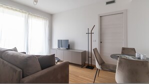 A modern, well-lit living room with a sofa, a TV on a cabinet, a dining table with chairs, and a door. - Italianway Comfort - Pichi 19 (Milan)