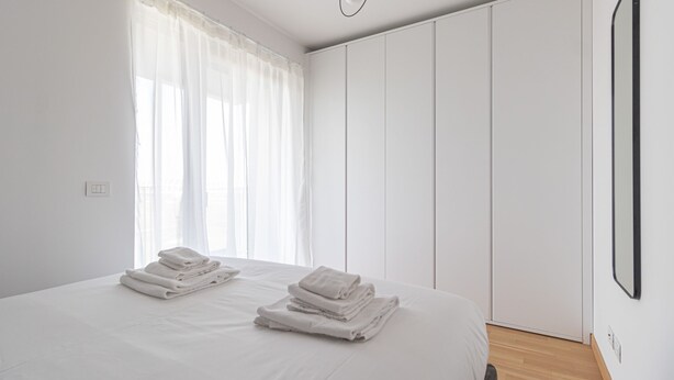 A bedroom with a bed, two sets of folded towels, a window with sheer curtains, and a wardrobe with vertical panels. - Italianway Comfort - Pichi 19 (Milan)