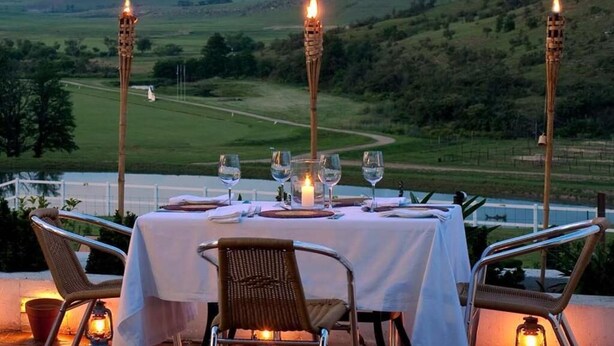 Ein Essbereich im Freien mit einem Tisch für zwei Personen, umgeben von Korbstühlen, mit Blick auf eine grüne Landschaft und Hügel im Hintergrund.