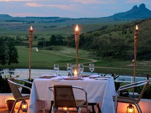 An outdoor dining setup with a table set for two, surrounded by wicker chairs, with a view of a green landscape and hills in the background. - Oaklands Farm Stay (Maluti a Phofung)