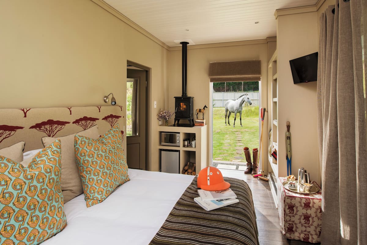 Un dormitorio con cama, almohadas estampadas, una mesita con un sombrero y vista a un caballo afuera.
