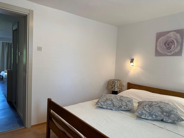 Une chambre avec un lit en bois, deux coussins à motifs, une lampe fixée au mur et une porte donnant sur une autre pièce.