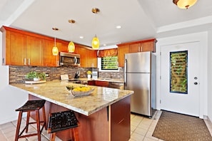 A modern kitchen with wooden cabinets, a stainless steel refrigerator, and a large island with stools. - Boutique Resort gem, spacious 1bd condo (Lahaina)