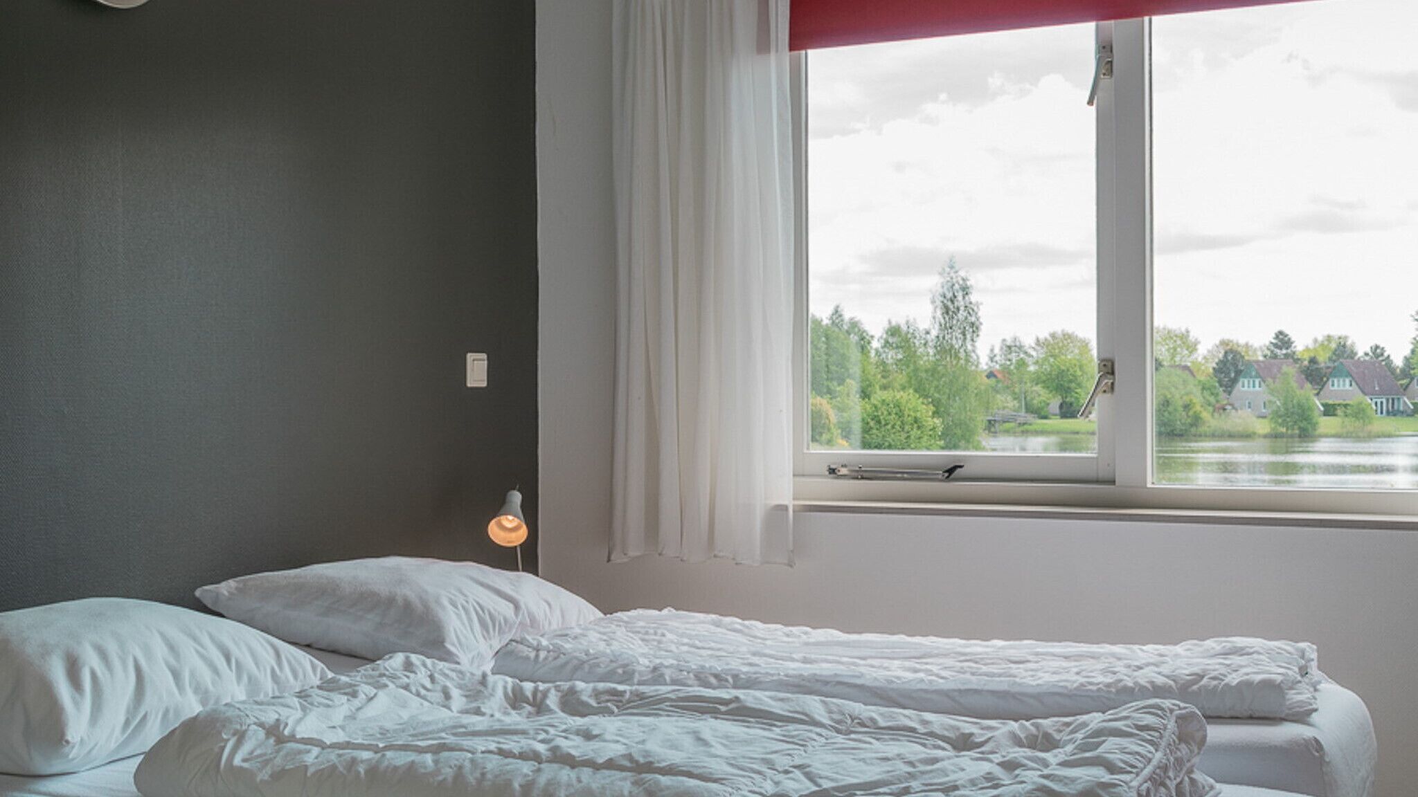 A bedroom with a bed, a window with a view of houses and trees, and a bedside lamp.