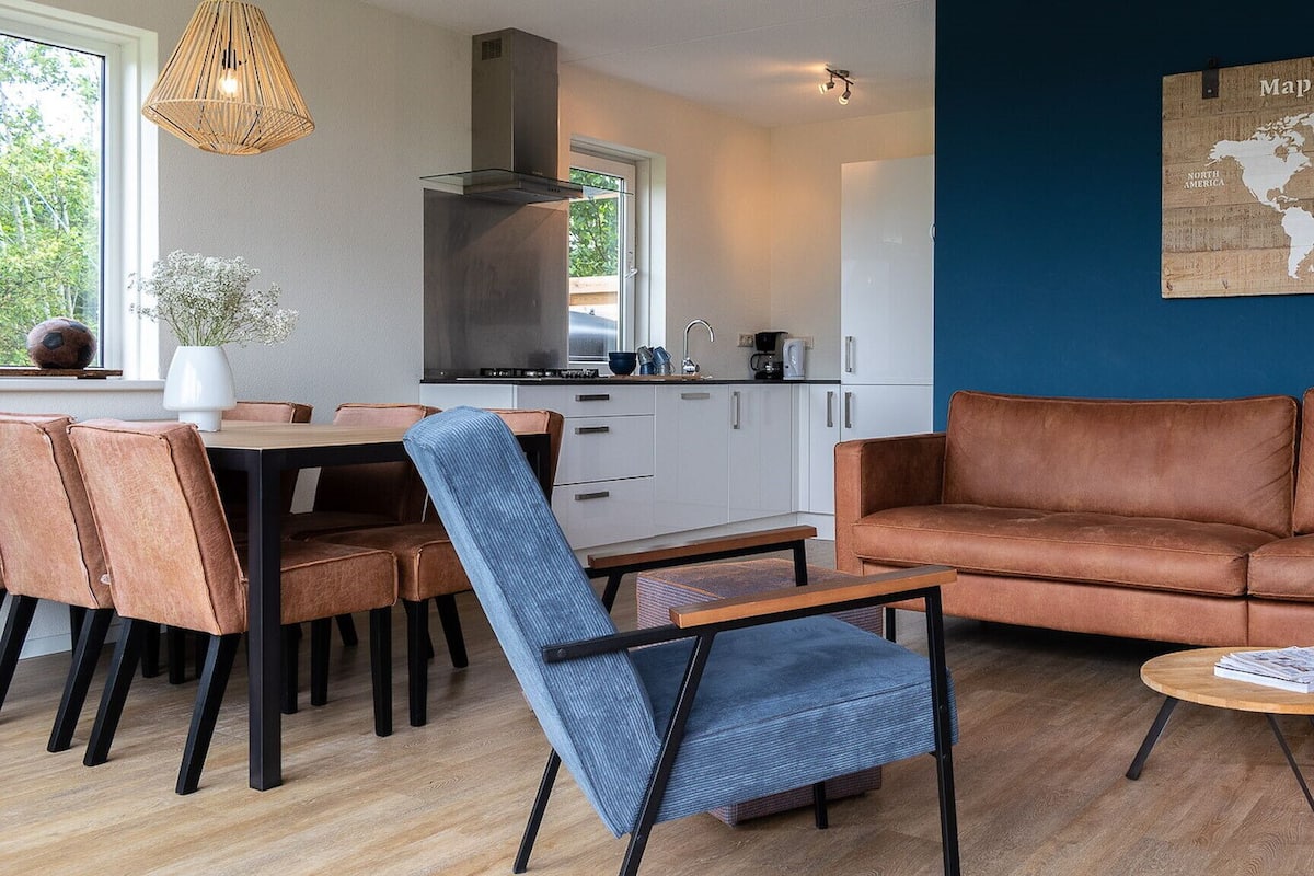 A modern living room with a brown leather sofa, a dining table with chairs, and a kitchen area with white cabinets and a stove.