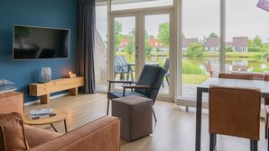 Una sala de estar moderna con un televisor grande, asientos cómodos, un banco de madera y vistas a un lago y casas.