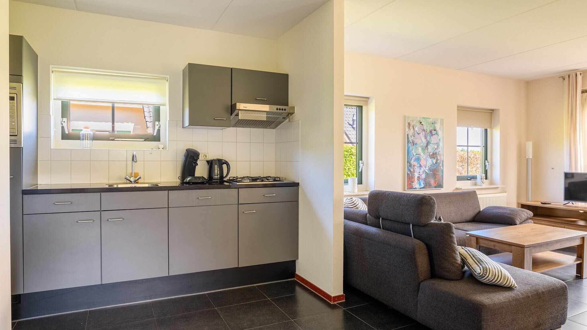 A modern kitchen with grey cabinets, a gas stove, and a coffee maker. There is a grey sofa and a wooden table in the living area.