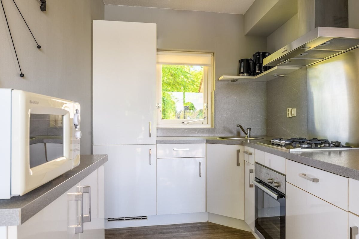 A modern kitchen with white cabinets, a built-in oven, a microwave, a gas stove, and a window with a view of greenery.