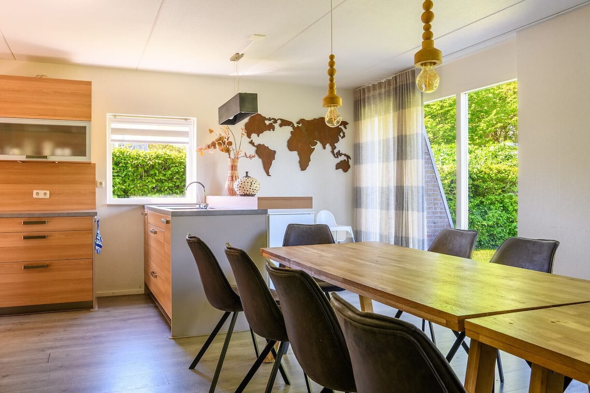 A modern kitchen with wooden cabinets, a dining area with a wooden table and chairs, a world map wall art, and a window with a view of greenery.