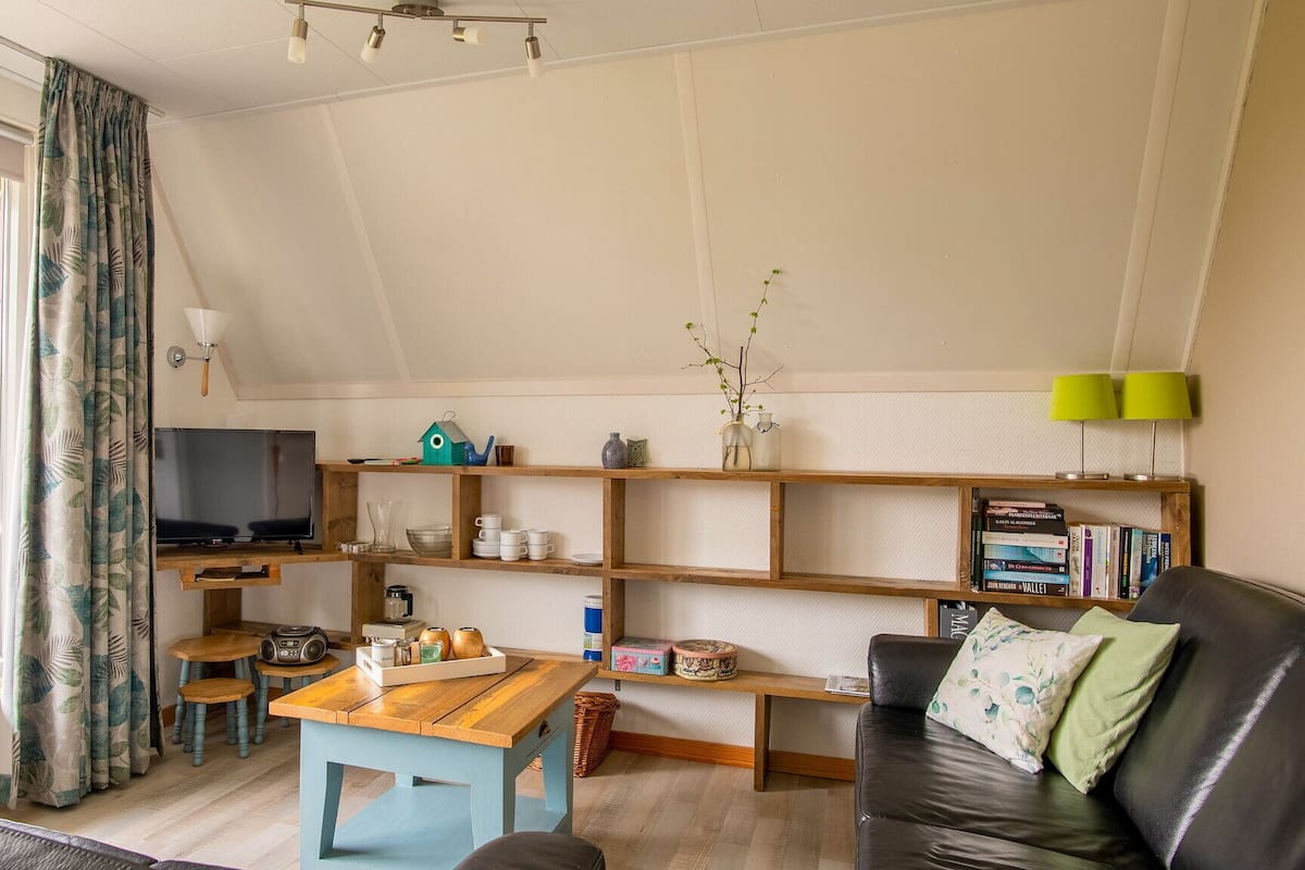 A cozy living room with a TV, wooden shelves, a sofa, and a window with curtains.