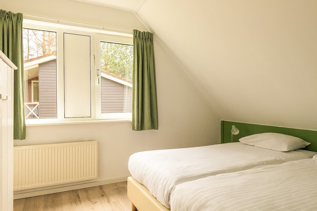 A bedroom with two beds, a window with curtains, a radiator, and a green headboard.