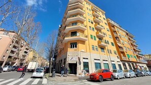 Un immeuble jaune à plusieurs étages, doté de balcons, avec une devanture portant l'inscription « IL BAZAR », et des voitures garées le long de la rue.