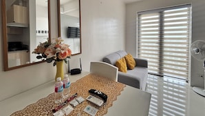 Un salon moderne avec un canapé blanc, une table basse ornée d’un chemin de table décoratif, un vase de fleurs, un téléviseur à écran plat et une fenêtre avec des stores.