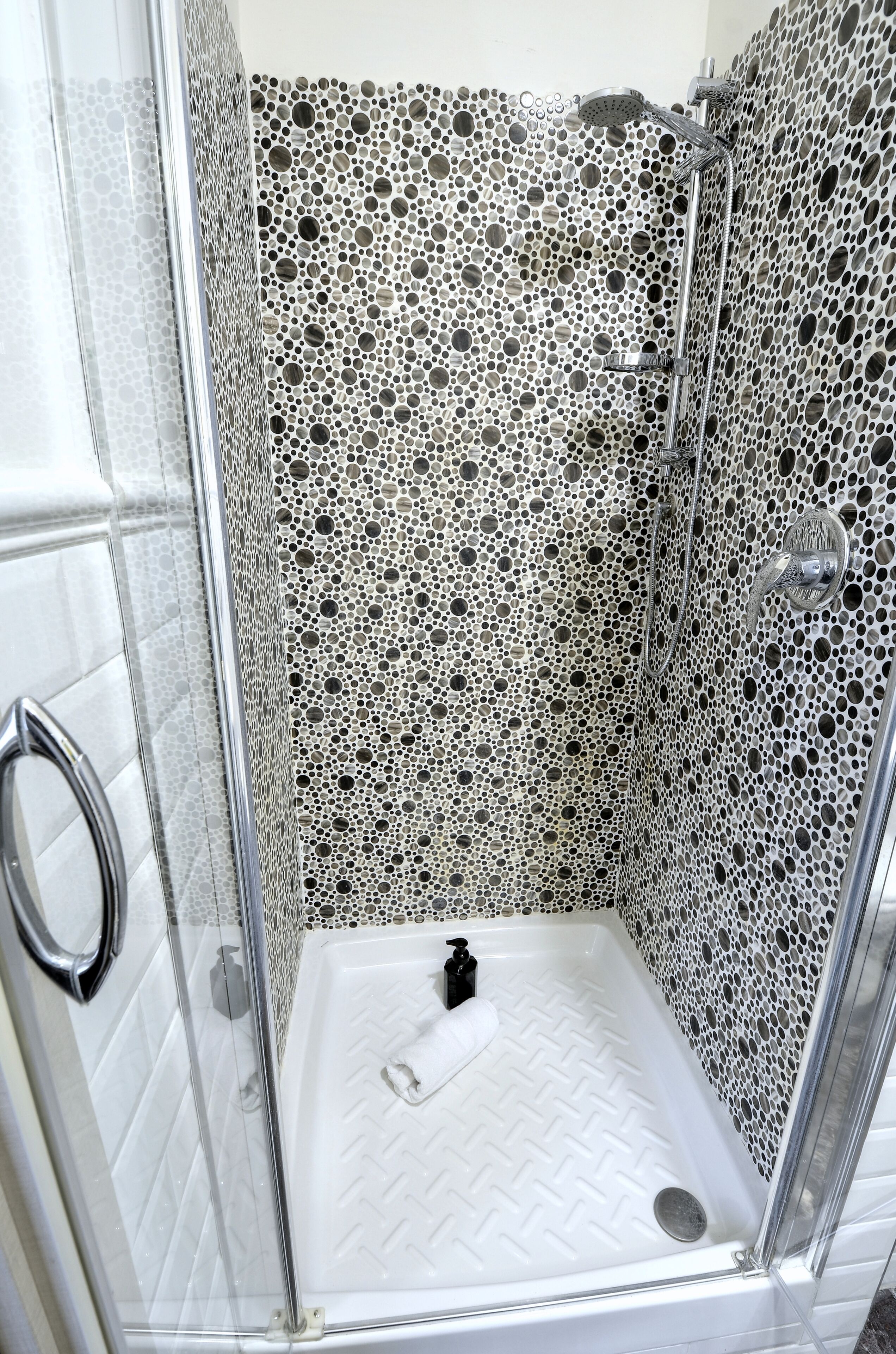 Um chuveiro com box de vidro, parede perfurada, chuveiro manual e piso branco.