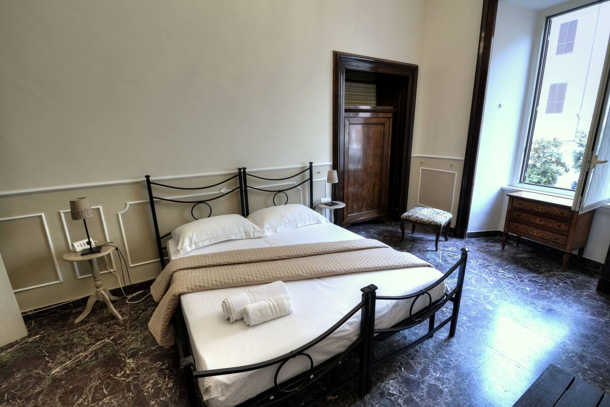 A neatly made bed with a white comforter and pillows, a bedside table with a lamp, a small wooden bench, and a window with a view of buildings.