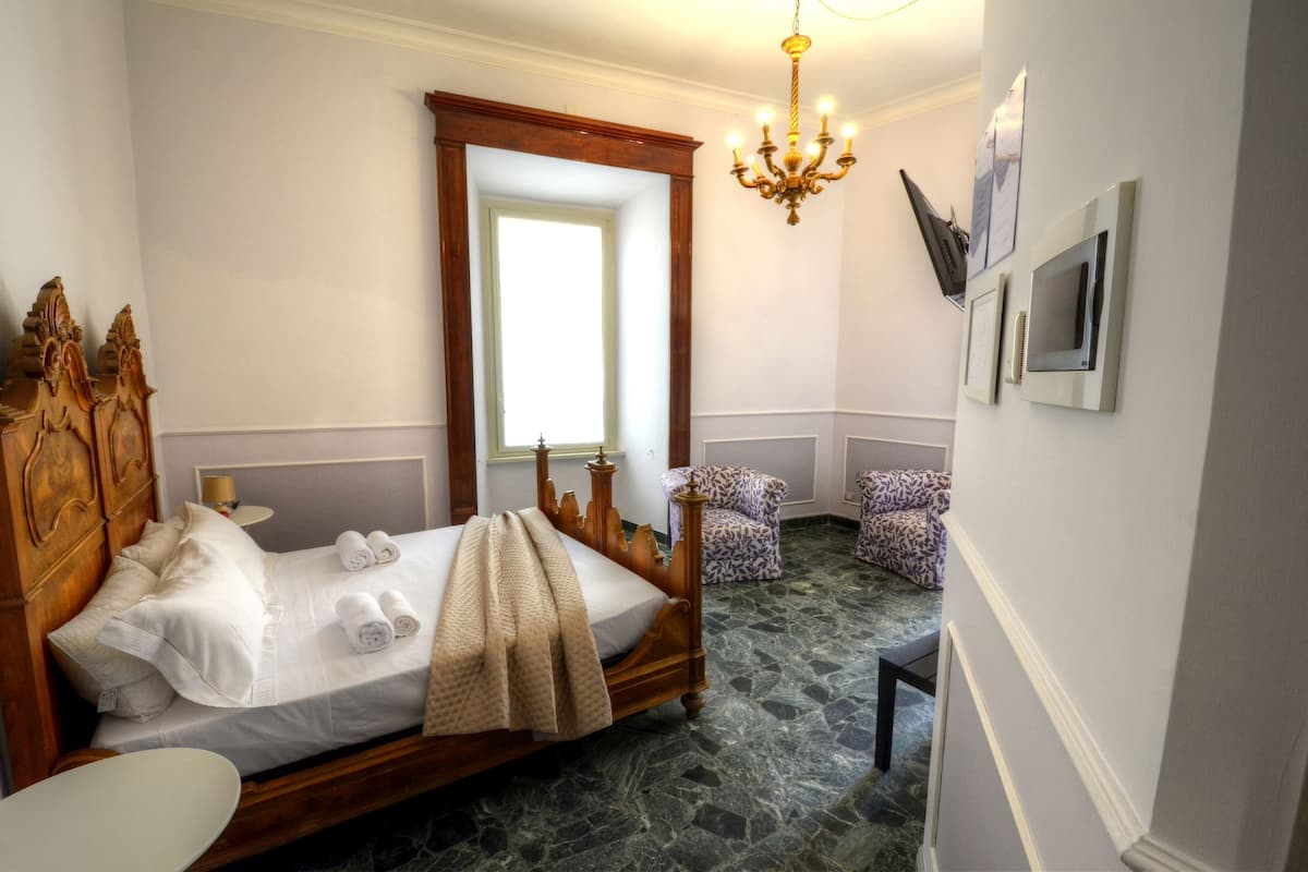 A bedroom with a wooden four-poster bed, a small round table, a patterned armchair, a chandelier, and a large mirror.