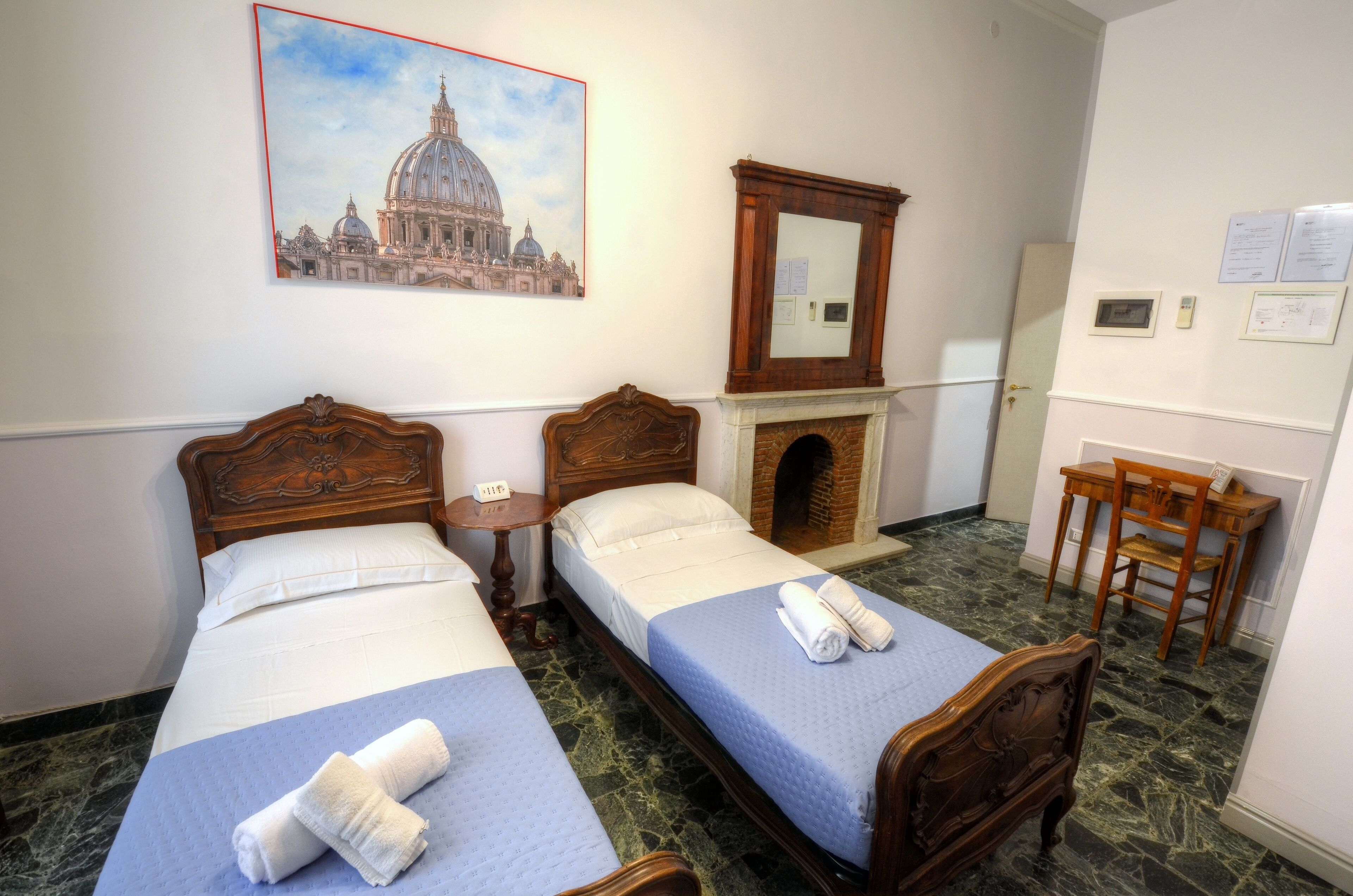 Een hotelkamer met twee bedden, een houten bureau, een stoel, een open haard en een schilderij van een historisch gebouw.
