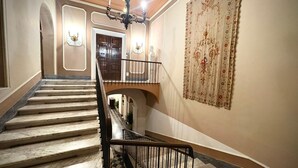 A grand staircase with marble steps and a black metal railing, leading to a landing with a chandelier and a door. - Palazzo Alibrandi (Civitavecchia)