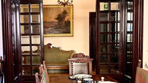 A dining room with a wooden table set for four, flanked by chairs with striped cushions. A painting hangs on the wall above the table. - Palazzo Alibrandi (Civitavecchia)