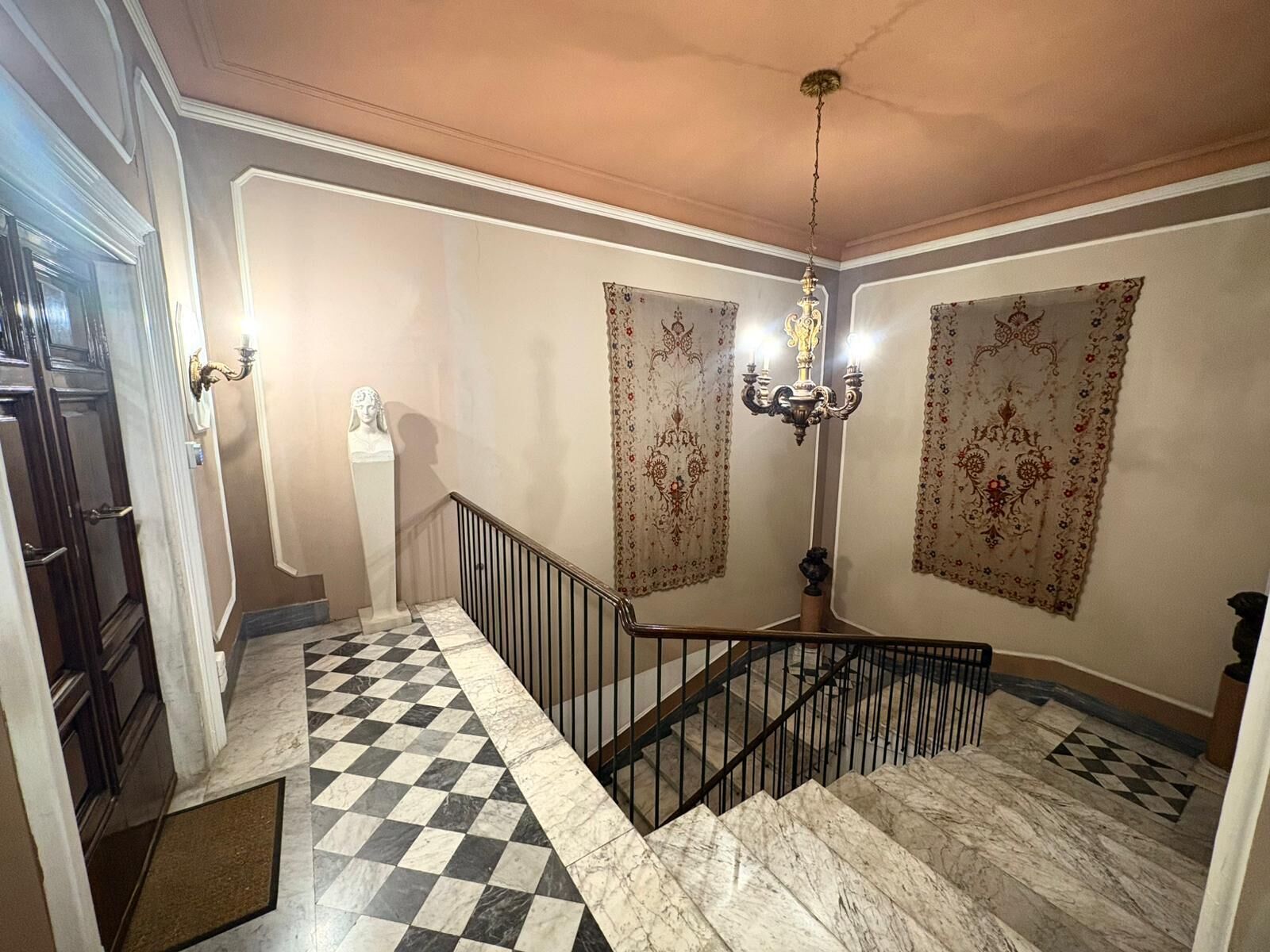 Un escalier avec un sol en marbre à carreaux, une rampe en métal noir et un lustre.
