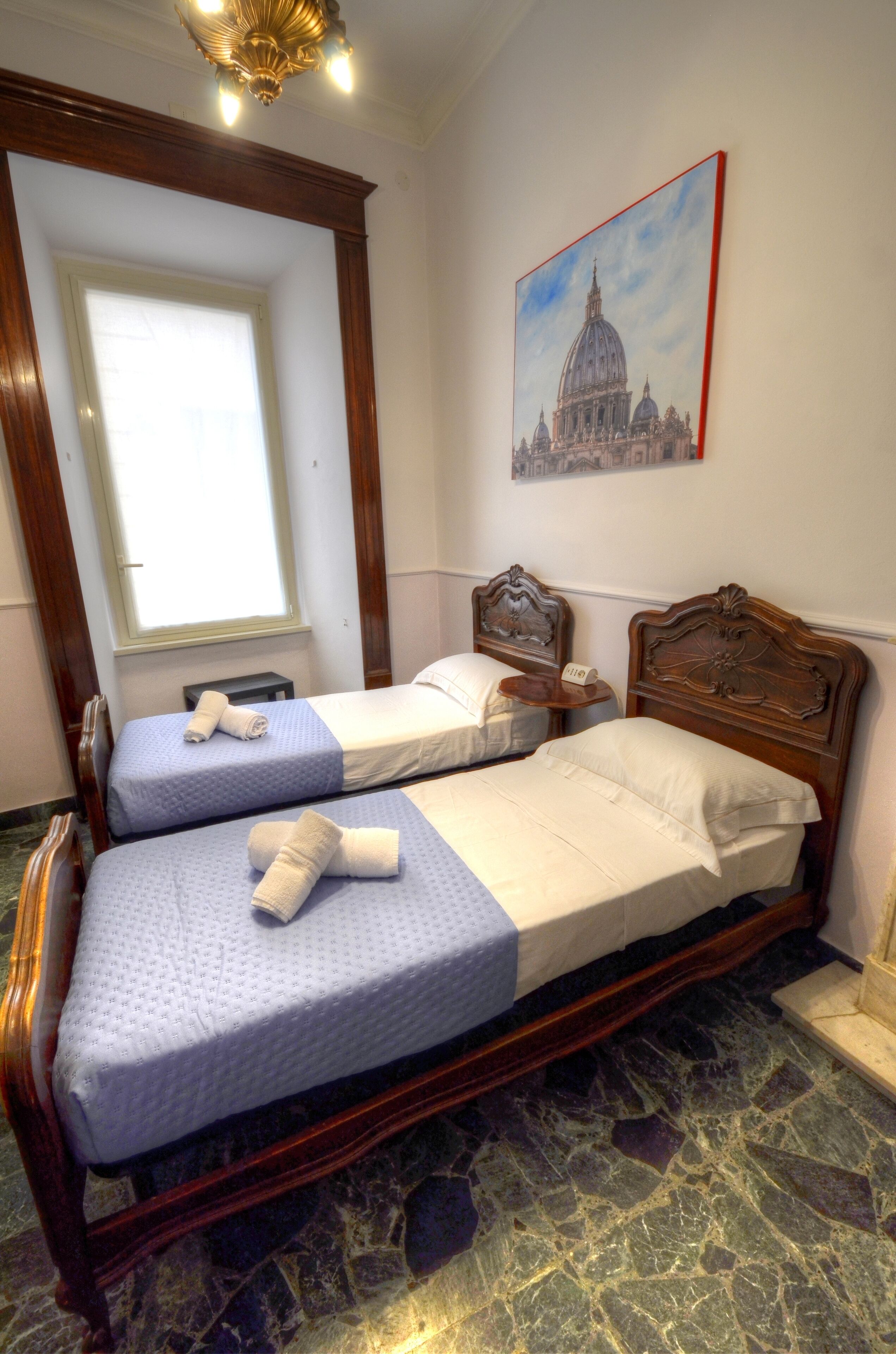 Une chambre avec deux lits, une fenêtre, un tableau représentant un bâtiment et un lustre.