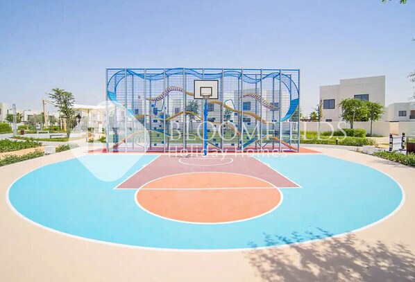 A basketball court with a blue and orange playing surface, surrounded by a fence with a basketball hoop.