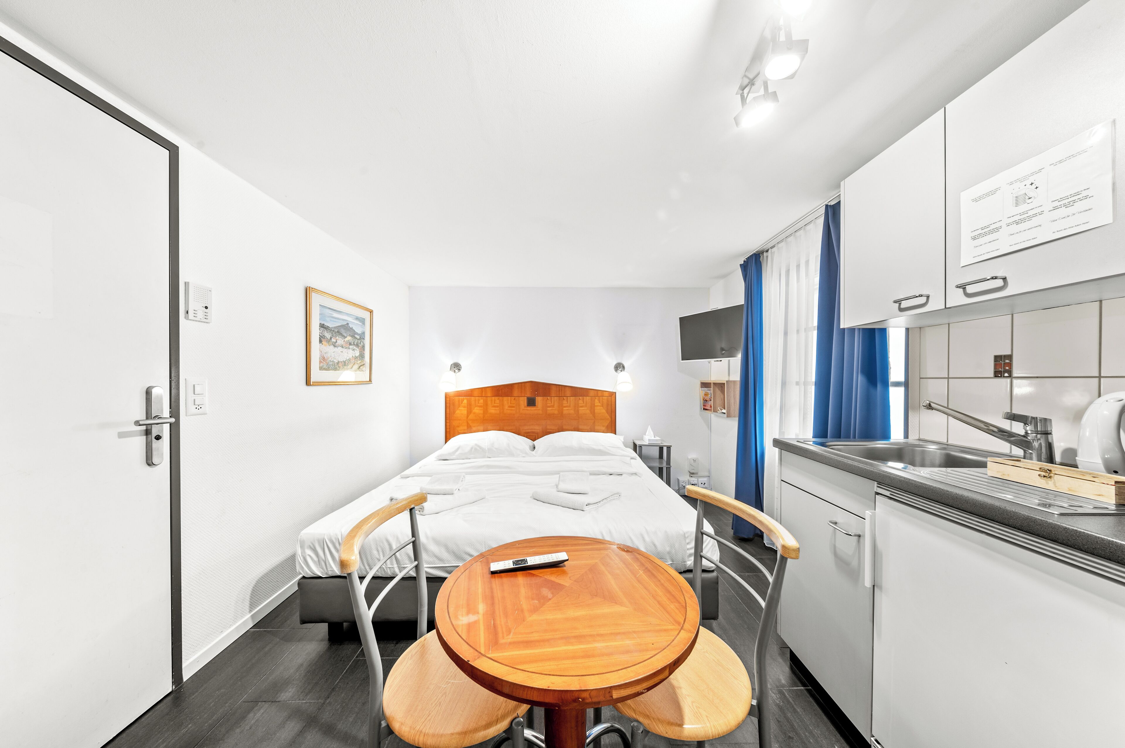 Habitación de hotel con cama, una mesita redonda con sillas, kitchenette y televisión en la pared.