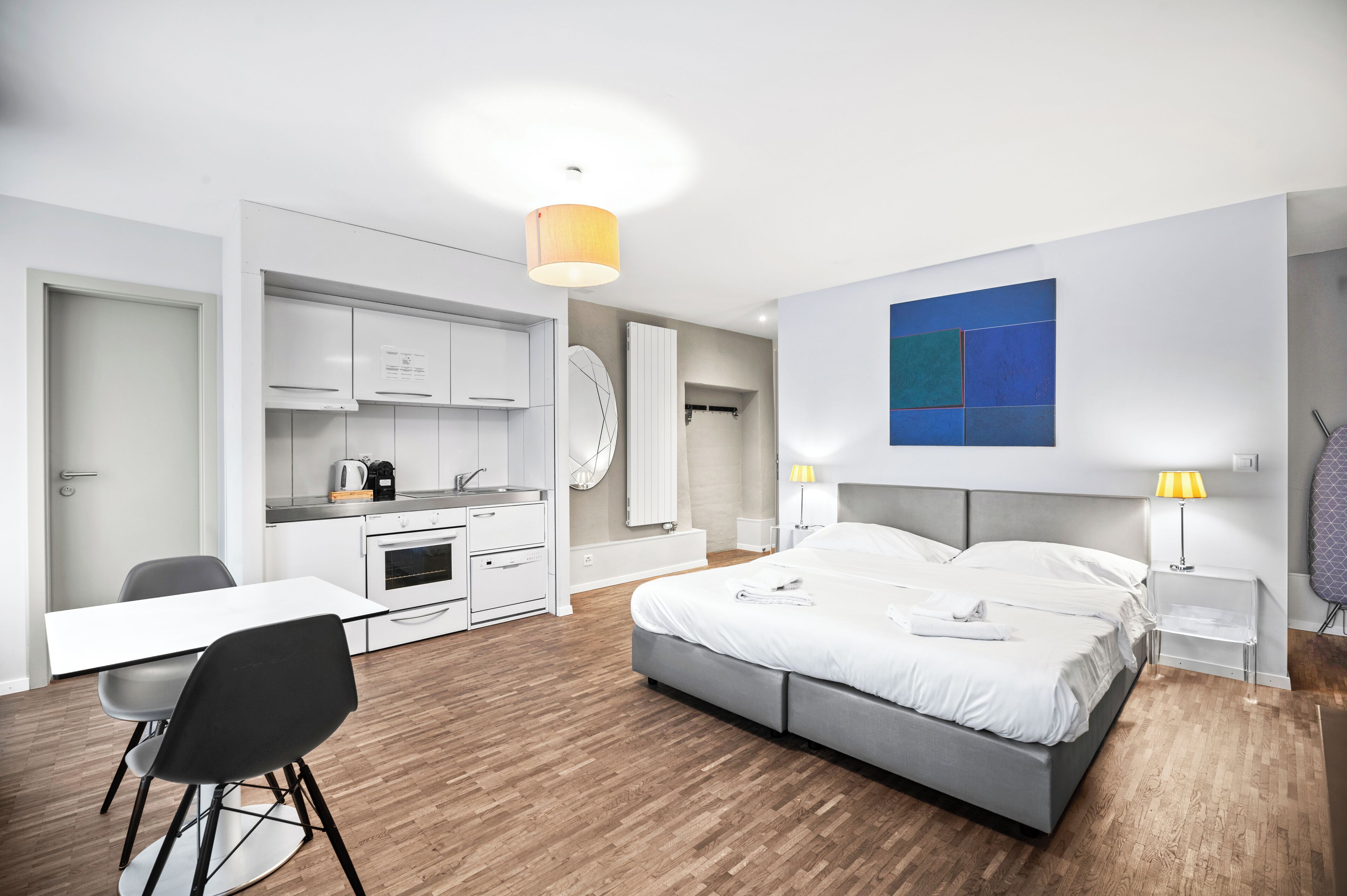 Una habitación de hotel moderna con cama, mesitas de noche, escritorio, silla, kitchenette y un amplio ventanal.