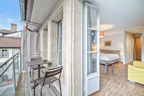 A balcony with a table and chairs, a bed with a white headboard, and a yellow armchair in a room with wooden flooring and large windows. -  Hine Adon Fribourg City Aparthotel Self checkin (Fribourg)