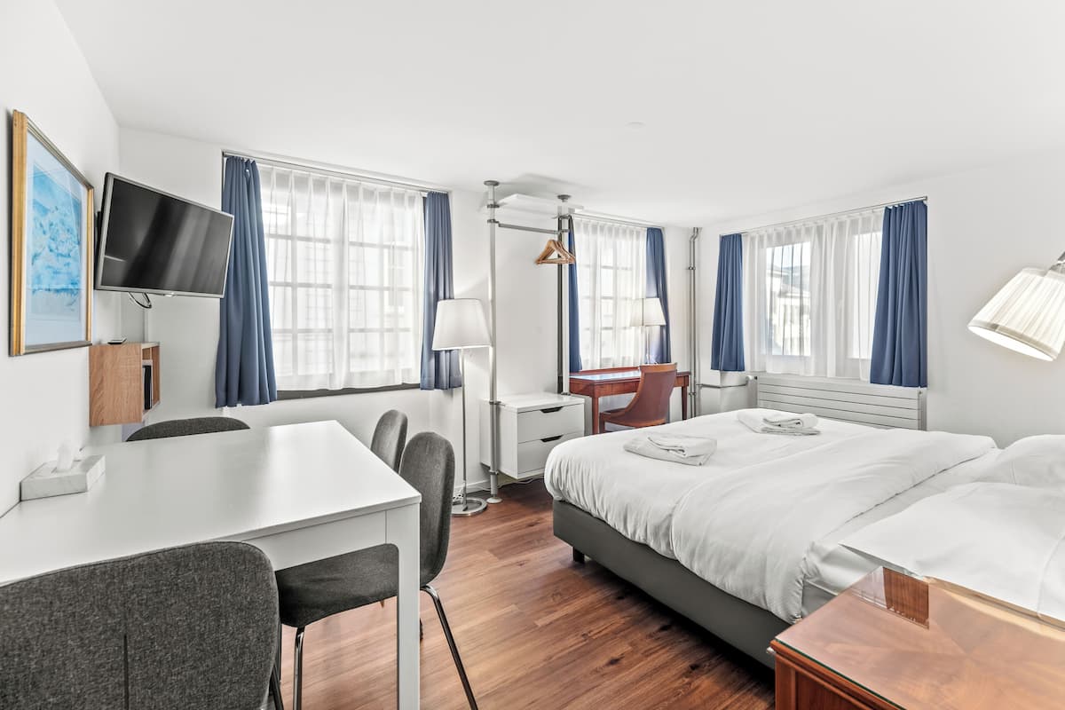 Ein Hotelzimmer mit Bett, Schreibtisch, Stuhl, Fernseher und Fenstern mit Vorhängen.