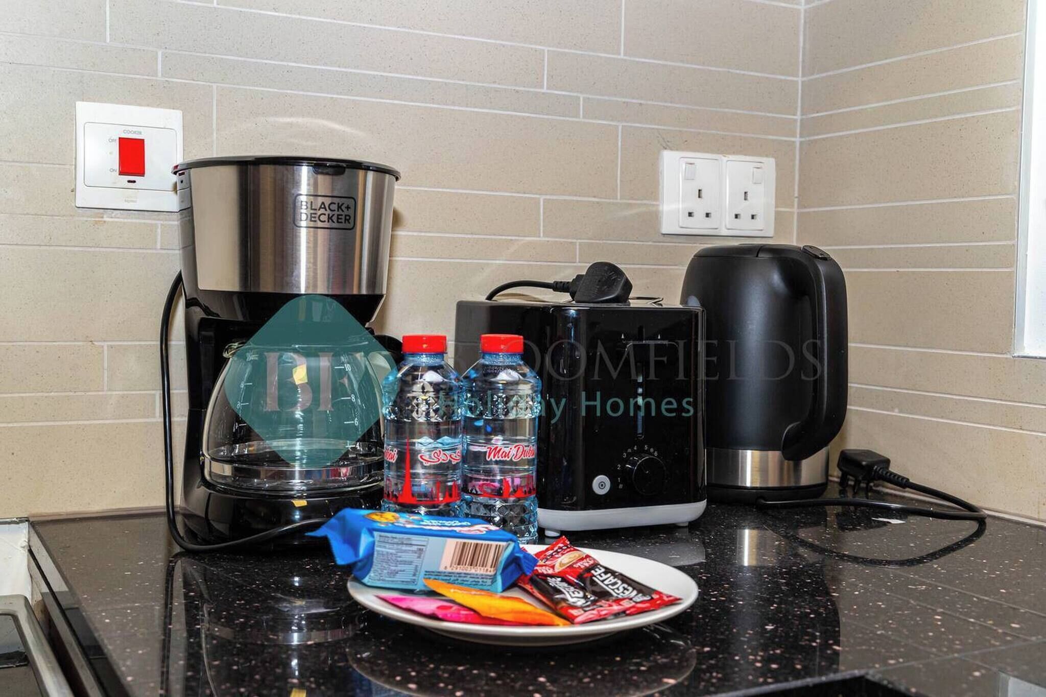 Una encimera de cocina con una cafetera, una licuadora y un tostador. Hay botellas de agua y un plato con aperitivos.