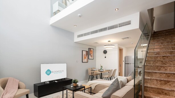 A modern living room with a black entertainment unit, beige sofa, and a staircase. - Bloomfields Premium 1BR Duplex in Rukan (Dubai)