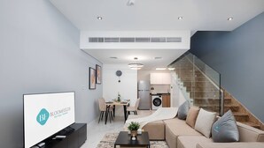 A modern living room with a sofa, coffee table, and TV. There is a dining area with chairs and a table. A kitchen with a fridge and washing machine is visible. A staircase leads to an upper level. - Bloomfields Premium 1BR Duplex in Rukan (Dubai)