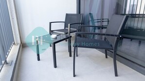 A balcony with a glass table and four chairs, a window with a view of buildings, and a metal railing. - Bloomfields Metropolitan 3BR In Bridges (Abu Dabi)
