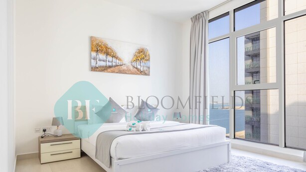 A modern bedroom with a large window, a bed with white bedding, a nightstand, and a painting on the wall. - Bloomfields Metropolitan 3BR In Bridges (Abu Dabi)